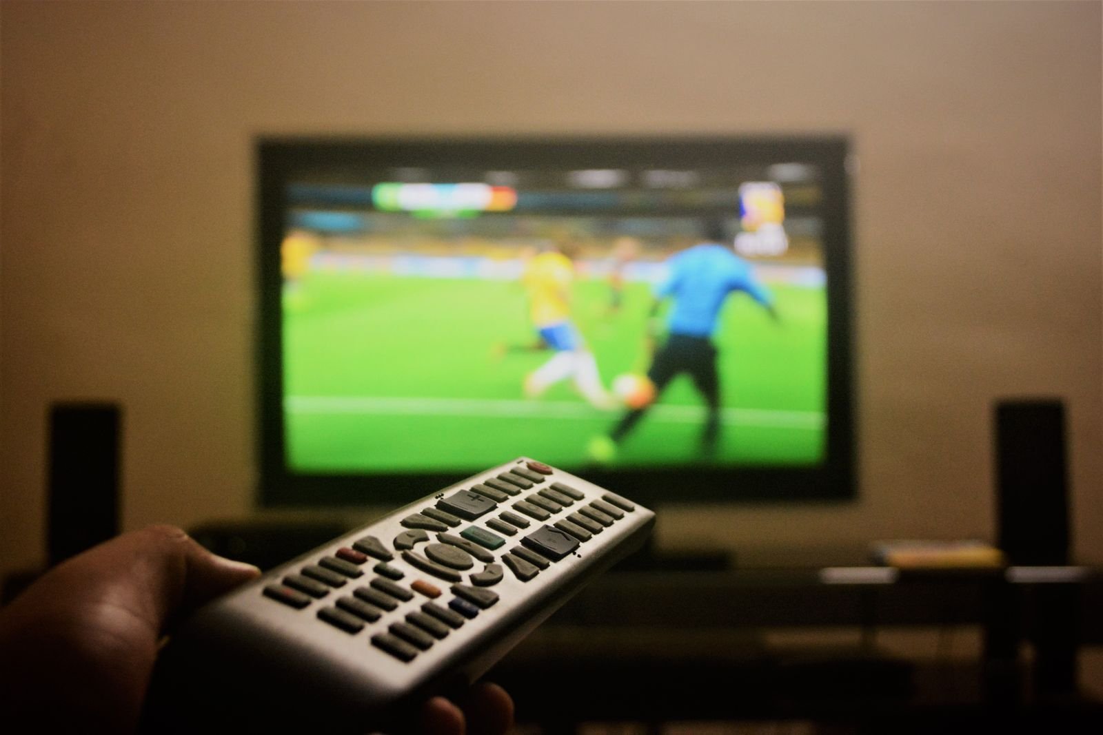 Hånd med fjernkontroll foran en tv som viser en grønn fotballbane
